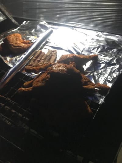 On a metal rack placed on aluminum foil, fried chicken pieces are arranged. Some pieces appear to be ribs, while others are bone-in chicken. Light reflects off the foil, revealing its crumpled surface. The setting is dim, with some areas obscured in darkness. A metallic tool rests near the chicken.