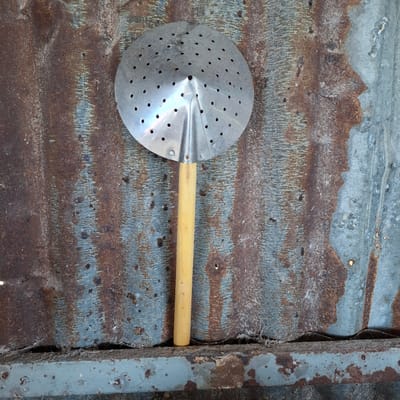 An overhead shot shows a metal strainer with a wooden handle resting against a corrugated metal surface that is heavily rusted and weathered. The strainer is round and has numerous small holes. The wooden handle is light in color and cylindrical. The corrugated metal background has a textured appearance with streaks of rust and faded blue paint. The bottom of the image shows a horizontal metal surface, also rusted and weathered, with some debris on top. The scene appears to be outdoors, possibly in an industrial or rural setting, and given the context of Rhieng Krueng, Indonesia, it might be in a backyard or a workshop.