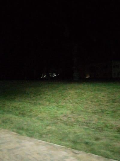 A dark, blurry image shows a dimly lit grassy field. The foreground features a concrete path. In the distance, faint lights indicate buildings obscured by the darkness. The overall atmosphere is eerie and indistinct due to the poor lighting and motion blur.