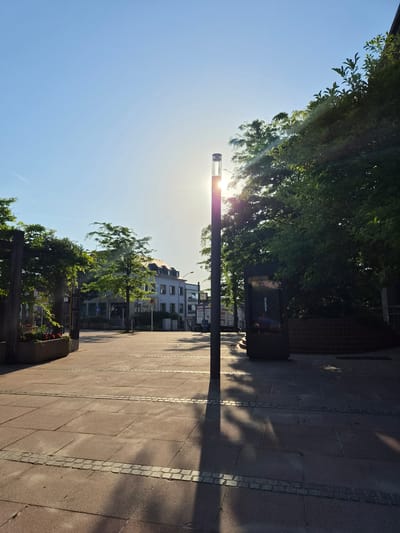 In the city of Sanem, Luxembourg, a street lamp stands prominently in the foreground of an outdoor plaza. The sun is positioned behind the lamp, creating a lens flare effect and casting a long shadow across the red-tiled ground. Lush green trees flank the scene, with sunlight filtering through their leaves, dappling the ground with shadows. In the background, buildings line the street, their architecture suggesting a European setting. The sky is a clear, bright blue, indicating a sunny day. The plaza appears to be a public space, possibly a town square or walkway, with plants in planters and a digital display visible on the right side.