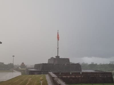 The image depicts the Citadel of Hue, a historical fortification located in Phú Xuân Ward, Vietnam. The scene is overcast and appears to be a gloomy day, possibly raining, with a thick haze in the background.

Dominating the center of the frame is the Flag Tower of Hue, a prominent structure within the Citadel. A tall flagpole extends upwards, topped by a Vietnamese flag, which is red with a yellow star. The tower itself is a substantial, multi-tiered stone structure with crenellated battlements.

The Citadel walls are visible in the foreground and extending to the right, constructed from dark stone. These walls are robust and imposing, suggesting a historical military significance.

To the left of the Citadel, a wide, paved area with green grass borders it. A lamppost stands prominently on this paved area. Further in the background, through the haze, other structures can be seen, including a smaller, ornate building with a curved roof that is reddish-orange in color. Trees and a distant cityscape are also discernible through the atmospheric conditions. The overall impression is one of historical grandeur under a somber sky.