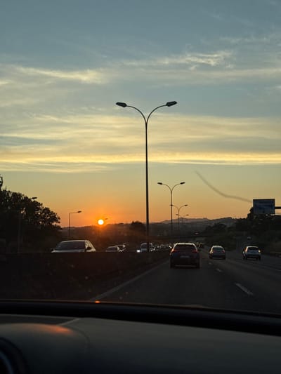 The scene depicts a multi-lane highway at sunset in Amadora, Portugal, observed from the interior of a vehicle. The sun, a bright orange orb, is low on the horizon, positioned left of the frame's center, casting a strong backlight. The sky above transitions from light blue to a gradient of yellow and orange near the horizon, featuring wispy cirrus clouds. A distinct dark, bifurcated streetlight pole stands prominently in the mid-ground, with a series of shorter, similar streetlights receding along the highway's central median.

Traffic is present on the highway; a dark sedan is directly ahead of the camera vehicle in the center lane, with additional vehicles visible in both forward lanes and on a parallel road to the left, separated by a concrete barrier. Many vehicles show illuminated headlights or taillights. Distant hills and structures are silhouetted against the horizon. On the far right, a highway sign, partially obscured, displays the word "AMADORA" on a blue background. A faint, dark, curved streak, consistent with a contrail, is visible in the upper right quadrant of the sky. The lower portion of the image shows the dark dashboard of the camera vehicle.
