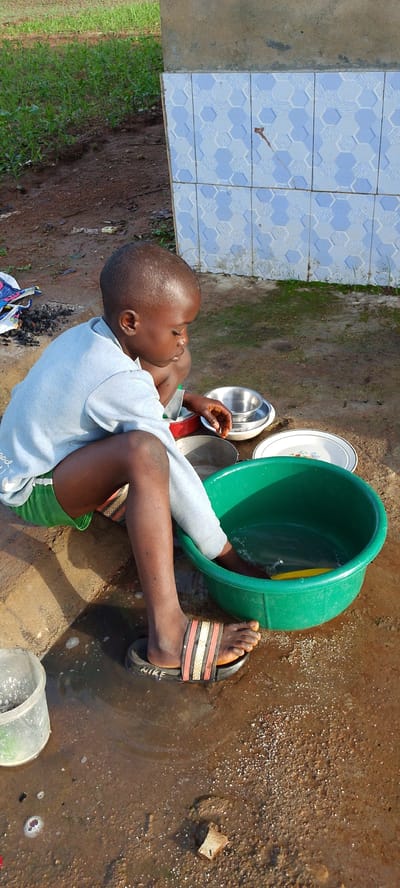 A young child is crouched down on the ground, with a large green basin of water in front of them. The child is wearing a light blue long-sleeved shirt and green shorts. Their right foot is in the water, and they appear to be washing something. Several metal and ceramic bowls are placed around the basin. The ground is dirt and appears wet, with some puddles. In the background, there is a wall with blue and white hexagonal tiles and some green grass. A clear plastic container is visible to the left of the child. The child is wearing black slides with three colored stripes and the word "NIKE" on the strap.
The scene suggests a task being performed outdoors, possibly in a rural or less developed setting, given the simple setup and the natural environment. The lighting indicates it is daytime. The presence of washing bowls and water suggests a chore or preparation activity. There is no clear indication of the specific time of day beyond it being daylight. The weather appears to be warm enough for outdoor activities, and there is no indication of rain. The location context of Gumel, Nigeria, suggests a possible West African setting. The child's expression is one of concentration on the task at hand.