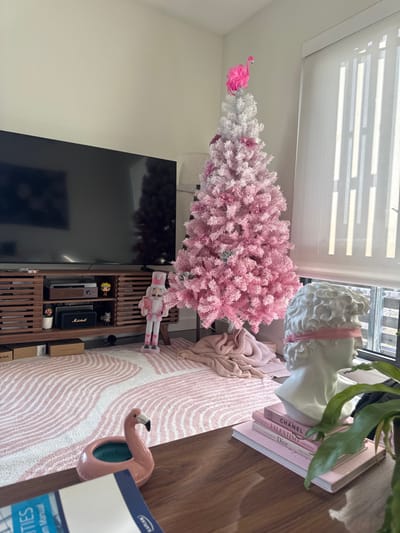 A pink and white Christmas tree stands next to a TV and a wooden media console. A pink flamingo ornament adorns the tree's top. A nutcracker figurine rests on the media console. A white bust statue with a pink ribbon sits atop pink fashion books on a wooden surface. A pink and white striped rug lies on the floor, where another flamingo object and an instruction manual can be seen. Blinds cover the window.