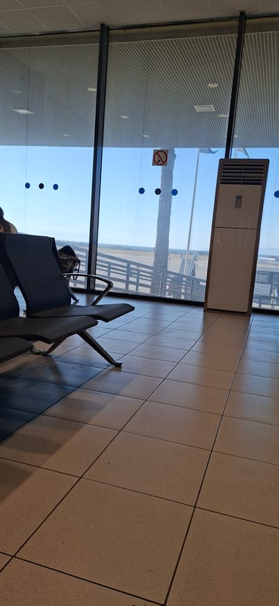 This media file displays an indoor scene, likely an airport waiting area, with a large window overlooking an outdoor environment. The predominant colors are the light brown of the tiled floor and the bright blue of the sky.
Inside, several dark gray chairs are arranged in rows. One chair is partially occupied by a person wearing dark clothing. Luggage, a dark backpack, is placed on a seat. To the left of the chairs, a tall, light-colored vertical air conditioning unit is positioned near the window. A round sticker with a red circle and a diagonal line through a cigarette, indicating "no smoking," is visible on the window. There are also several small blue circular decals on the window.
Through the window, an airport tarmac is visible with what appears to be part of an aircraft and some railings. The sky is clear and bright blue, suggesting it is daytime, likely a sunny day. The overall atmosphere is one of waiting and travel.
The location is specified as Rhodes, Greece, which aligns with the bright, sunny weather depicted. No discernable text is visible in the image apart from the "no smoking" symbol.