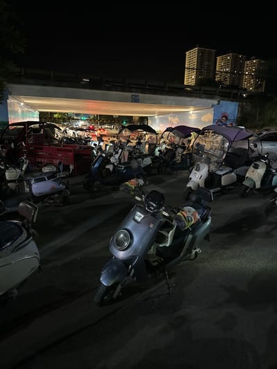At night in Eshan Yi Autonomous County, China, a variety of electric scooters are parked on a paved surface beneath an overpass. The overpass's underside features a mural. In the background, several multi-story buildings are illuminated, and traffic lights are visible on a street beyond the overpass. The foreground shows a collection of electric scooters, some equipped with transparent protective covers. One scooter in the center has a grey body and a visible kickstand. To the left, a portion of a white scooter is visible. The lighting suggests a typical urban night scene.