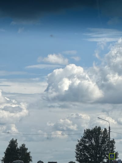 The image shows a cloudy sky above a street scene. In the foreground, there are trees and a few cars. A street light with a pedestrian crossing sign is visible on the right side of the image. The sky is filled with large, fluffy white clouds against a blue background. The overall impression is of an overcast day in a residential or suburban area.