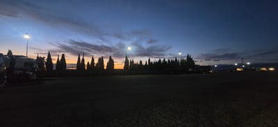 A wide panoramic image captures a parking lot at dusk in Épila, Spain. The sky is a deep blue, transitioning to orange and pink hues near the horizon, with scattered clouds displaying wispy textures. Several tall, dark cypress trees form a silhouette line across the middle ground, interspersed with what appear to be streetlights casting bright beams. In the foreground and mid-ground, multiple large trucks and their trailers are parked, silhouetted against the twilight sky. Some trucks have their headlights on, casting a warm glow. The ground is paved with asphalt, showing a textured surface. The overall scene suggests a quiet, post-sunset atmosphere in a truck stop or industrial area.