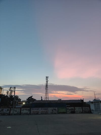 The image depicts a view of the sky and a city scene in Sitarganj, India, during what appears to be twilight or dawn. The sky is a gradient of colors, transitioning from a clear blue on the left to soft pinks and oranges on the right, indicative of sunset or sunrise. Several clouds are present, tinged with the colors of the sky.
Dominating the mid-ground is a tall, metallic telecommunications tower with multiple antennas. To the left of the tower, a cluster of trees and smaller structures are visible. Below these, a low building with a weathered facade runs across the frame. This building features graffiti and what appear to be advertisements or signs written in Hindi. One sign prominently displays "डेग मच्चर को भगला है जिन्दगी को बचाना है" (Deg machhar ko bhagala hai jindagi ko bachana hai), which translates to " Dengue mosquito needs to be driven away, life needs to be saved." Below this, smaller text reads "नगर पालिका परिषद" (Nagar Palika Parishad), indicating it is a municipal council notice.
To the right of the main building, another building with white walls and closed doors is partially visible. The foreground consists of a paved, open area, likely a road or a plaza, with visible seams in the concrete. The overall impression is an urban landscape at the edge of the day, with a focus on the sky, infrastructure, and local signage.