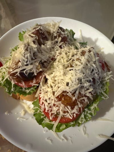 This is a close-up, overhead shot of two open-faced sandwiches on a white plate. Each sandwich is made with a slice of toasted bread, topped with lettuce, tomato slices, and a mixture of what appears to be ground meat or small meatballs, and olives. The sandwiches are generously covered with shredded white cheese. The lighting is dim, and the background is out of focus, showing a wall. The scene is indoors, likely in a residential setting in Peremyshliany, Ukraine.