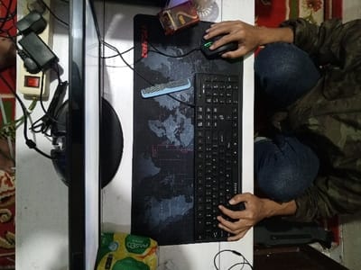 An overhead view depicts a computer workstation located in Al Haouafate, Morocco. A person, visible from the chest down, wears a camouflage-patterned upper garment and blue jeans, positioned in front of a white desk. The individual's right hand operates a black gaming mouse with green LED accents on a black mouse pad featuring a grey world map design. The left hand rests on a black full-size keyboard. To the left of the setup, a black computer monitor is visible from its side. Adjacent to the monitor, a white power strip with a red illuminated switch is connected to several black power adapters and cables. Other objects on the desk include a small blue and silver item (possibly a USB device), a partially visible brown box, a transparent cup with a metallic rim, and a yellow and green "PASEO Green" package. The floor beneath and to the left of the desk is covered by a patterned rug featuring red, green, and cream colors.