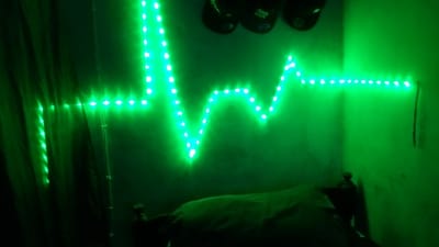An interior scene in Ndeiya, Kenya, illuminated by green LED strip lights affixed to a wall. The LED strip forms a horizontal, undulating pattern resembling an electrocardiogram waveform. Above the LED strip, three dark, indistinct objects are partially visible, possibly hanging from the upper wall or ceiling. Below the LED strip, a dark-colored bed with a visible headboard and a rumpled cover is positioned. The room's walls are bathed in a prominent green hue originating from the LEDs. No individuals are present in the image.