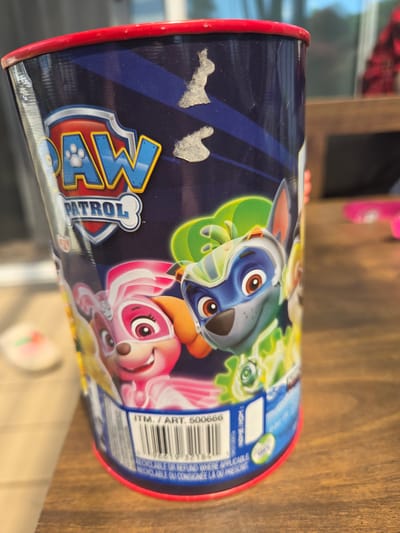 The image displays a dark blue cylindrical container, likely a can or cup, from the "Paw Patrol" franchise, resting on a wooden surface. The container features vibrant illustrations of several Paw Patrol characters, including what appear to be Chase (blue/grey) and Skye (pink), in a stylized, superhero-themed design with neon green and yellow accents. The iconic "PAW PATROL" logo, designed as a shield with "PAW" and a bone shape with "PATROL," is prominently displayed on the left side of the container.
Two light-colored, irregularly shaped smudges are visible on the dark blue background to the left of the logo. The container has red rims at both the top and bottom. On the right side, there is a white label containing a barcode and various pieces of text. Visible text includes "ITM. / ART. 500666," "NAME/NOM," and "19KS130678." The barcode numbers "966191321844" are also present. Additionally, text indicating recyclability is seen: "RECYCLABLE OR REFUND WHERE APPLICABLE" and "RECYCLABLE OU CONSIGNEE LA OU PRESCRIT." The lighting suggests an indoor setting, with some reflections on the container's surface. No specific activity or people are depicted; the image focuses solely on the Paw Patrol container.