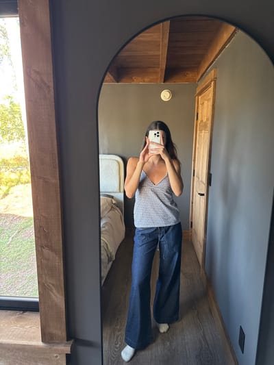 An adult female subject is captured in a mirror selfie within an indoor room. She wears a blue and white gingham-patterned spaghetti strap top, dark blue wide-leg drawstring pants, and white socks. Her upper face is obscured by a light-colored smartphone, held in both hands, which features a triple-lens camera array. Long, dark hair frames her face, and a silver-colored necklace with a small pendant is visible.
The full-length, arch-top mirror with a thin black frame reflects the room's interior. To the subject's left, a bed with a light-colored, possibly upholstered headboard and light-colored bedding is visible. To her right, a light-colored wooden door with a dark handle is set into a dark grey wall. A round, off-white smoke or carbon monoxide detector is mounted on the wall above the door. The ceiling features exposed wooden beams. The floor is constructed of light-colored wood planks. A large window, framed by dark wood, is partially visible on the far left, reflecting bright outdoor light and green foliage, indicating a daytime setting. The specific location within Otego, United States, cannot be determined beyond an interior residential or lodging space.