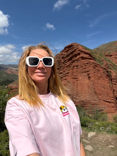 A woman with long blonde hair and white square-framed sunglasses smiles directly at the camera in a vertical, outdoor portrait. She is wearing a light pink t-shirt with a cartoon panda logo on the left chest, which also features the text "Panda HOLA!". The setting appears to be a sunny day, with a clear blue sky and some scattered white clouds visible in the upper and lower portions of the image.
Behind the woman, a dramatic landscape unfolds. Dominating the mid-ground are striking red-brown rock formations, characterized by their layered, almost striated texture, indicative of erosion. These mountains are partially covered with sparse green vegetation in certain areas. To the right of the woman, a dirt path or natural ground is visible, alongside more green bushes and small rocks. The overall impression is that she is standing in a mountainous or canyon-like environment. The image is shot from a slightly low angle, emphasizing the vastness of the sky and the height of the rock formations. Her expression is joyful and relaxed. Given the context, this scene is likely captured in Jeti-Ögüz, Kyrgyzstan.