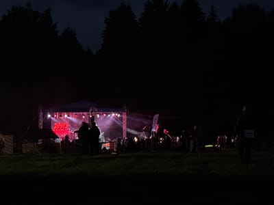 This is a nighttime photograph depicting an outdoor music event in Большая Кивара, Russia. The primary focal point is a brightly lit stage in the mid-left, adorned with multiple spotlights emitting pink and purple light. A large red neon-style sign on the stage clearly displays the text "DYMAT BEATS". Several indistinct figures are visible on the stage, likely performers or crew. To the left of the stage, there is a tent or canopy structure.
In front of the stage, a moderate crowd of people is gathered, their forms largely silhouetted against the stage lights, indicating they are watching the performance. Two individuals are more prominent in the mid-ground left, standing closer to the stage. The foreground consists of a dark grassy field.
Along the upper half of the image, a dense line of dark, silhouetted evergreen trees forms the backdrop, meeting a dark, cloudy night sky. On the far right in the foreground, a single individual is visible from behind, walking away from the stage. This person is wearing what appears to be a backpack or a numbered bib, with some faint light-colored markings on it. The overall illumination of the scene is dim, with artificial lights from the stage providing the main source of light. The image quality exhibits graininess consistent with low-light photography.