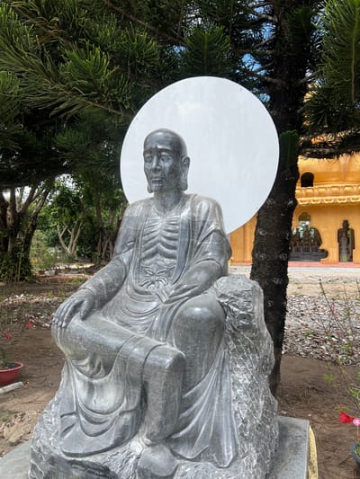 A dark grey stone statue depicting an emaciated, seated figure is centrally positioned outdoors in Đăk Mar, Vietnam. The figure possesses a bald head, elongated earlobes, a visibly open mouth, and a gaunt torso with distinct ribs sculpted beneath draped robes. Its hands rest on its lap, and it is seated on a textured, rock-like base. A large, circular white panel functions as a halo directly behind the figure's head. To the right of the statue stands a large tree with dark, textured bark and dense green foliage. To the left and in the background, additional green trees and vegetation are visible. The ground immediately around the statue consists of bare earth with scattered small stones and sparse red-flowered plants in small pots or bushes. In the background to the right, a multi-tiered building with golden-orange or yellow walls and arched openings is visible, indicative of a temple or pagoda structure. The sky above is partially visible, showing a light blue with some cloud cover.