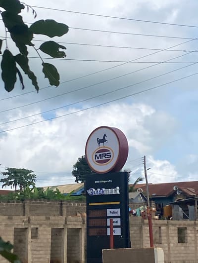 The image, rotated 90 degrees clockwise, captures an outdoor scene featuring a gas station or convenience store sign alongside buildings, power lines, and foliage under an overcast sky.
The central focus is a prominent sign structure. On the left is a circular sign displaying a dark silhouette of an animal, possibly a horse or goat, within a gold-bordered oval. Inside the oval, the letters "MRS" are clearly visible. Below this, smaller text reads "MRS Oil Nigeria Plc." To the right of the circular sign is a rectangular, dark-colored vertical sign with "QuickMart" written horizontally near the top in white and yellow text. Below "QuickMart," there are rectangular panels listing fuel types: "Petrol," "Diesel," and a partially visible label beginning with "G."
In the background, various buildings are visible. Some appear to be concrete structures, possibly residential or commercial, with unfinished elements like exposed blockwork, suggesting construction or renovation. Trees and other green foliage are scattered throughout the mid-ground. Numerous power lines stretch across the upper portion of the frame against a cloudy, bright sky, indicating daytime. Dark-silhouetted leaves are visible in the bottom-left foreground. No people or specific activities are discernible in the image. The scene suggests a roadside or urban environment, consistent with the provided location context of Sasaro, Nigeria.