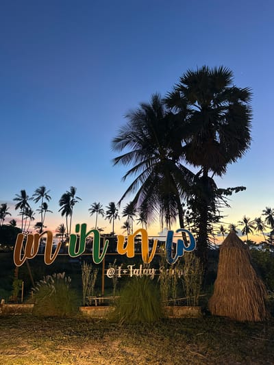 The image depicts a scenic view at dusk in Baan Taling Ngam, Thailand, characterized by a twilight sky transitioning from deep blue to soft orange and pink hues. In the foreground, a large, colorful sign with stylized lettering in brown, green, blue, and yellow stands prominently. Beneath the main sign, smaller text reads "@ I + Talay Samui." The sign is surrounded by lush green foliage, including tall grasses and what appear to be potted plants. To the right of the sign, a conical structure made of dried palm fronds, resembling a haystack or a traditional hut, adds to the tropical ambiance.

In the background, a dense arrangement of palm trees stretches across the horizon, their silhouettes stark against the vibrant sky. The diverse heights and shapes of the palm trees suggest a natural, untamed landscape. The overall scene conveys a sense of tranquility and tropical allure, likely representing a beachfront location or a resort area in Baan Taling Ngam. No people are visible in the image.