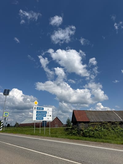 The image captures a rural roadside scene under a bright blue sky with scattered white clouds. A paved road runs diagonally across the foreground, with a white lane divider. On the left side of the road, a signpost displays destinations in Russian: "НЕРДВА 22", "СРЕТЕНСКОЕ 22", "ИЛЬИНСКИЙ 29", and "ЧЕРМОЗ 78". A yellow diamond-shaped sign is mounted above these destination signs, and a pedestrian crossing sign is visible to the left of the destination signpost. A solar panel is mounted on a pole near the signs.
In the background, several old buildings are visible. One building on the right has a dark wooden facade and a rusty corrugated metal roof. Another building behind it is a large barn with a corrugated metal roof and walls. Green grass and vegetation cover the ground around the buildings and along the roadside. The environment appears to be a rural area, possibly in or near the city of Рождественск, Russia, given the road signs. No people are present in the image.