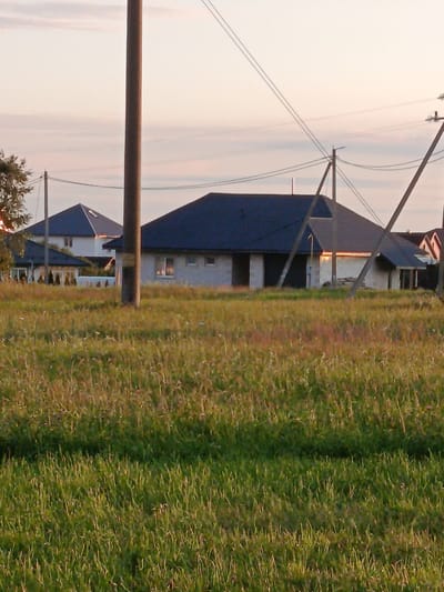 The image depicts a rural landscape in Смілавіцкі сельскі Савет, Belarus, during what appears to be sunset. A tall, brown utility pole stands prominently in the foreground, slightly off-center to the left. In the middle ground, several houses with dark, pitched roofs are visible. One house, closer to the center, is under construction with visible brickwork and window openings. Power lines crisscross the sky above the houses, supported by additional utility poles. The foreground is dominated by a field of tall, green and brown grass, indicative of a meadow or pasture. The sky above is a pale, hazy orange and white, suggesting the setting sun.