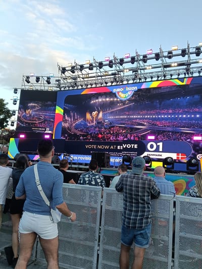 The image shows a crowd of people watching a large outdoor screen displaying the Eurovision Pride Maspalomas Festival in San Bartolomé de Tirajana, Spain. The screen shows a performance with the number "01" and the word "Denmark." Lighting rigs are visible above the screen. The crowd is standing behind metal barricades. Several people are visible, including a man with purple hair wearing a blue patterned shirt and white shorts, and another man in a plaid shirt and denim shorts.