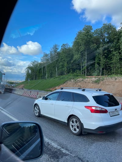 The image shows a view from a car traveling on a road in Novodrozhzhino, Russia. A white car is visible in the adjacent lane. The landscape includes trees, grass, and a blue sky with clouds. There are concrete barriers along the side of the road. The side mirror of the car taking the picture is also visible. The number plate of the white car is A080EA 105 RUS.