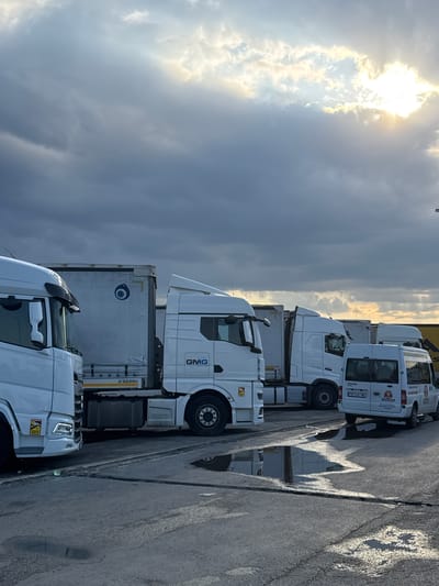 This image displays a row of parked white trucks, primarily semi-trucks and a few smaller vans, under a dramatic sky with sunlight breaking through dark, overcast clouds. The sun appears to be setting or rising, casting a warm, diffused glow and creating highlights on the wet asphalt ground, suggesting recent rain. The trucks are parked on a paved surface, likely a lot or a rest stop.
Several trucks have visible logos and text. One semi-truck has "GMG" in large blue letters on its side. Another truck's trailer has a circular logo with a blue eye-like design and the text "SERIN". A smaller white van has "TRANSIT" and the license plate "PB 0341 ME". There is also a yellow warning sticker on the front bumper of one of the semi-trucks.
The overall mood is calm and still, with the focus on the vehicles and the atmospheric sky. The lighting suggests it is either early morning or late afternoon. The presence of multiple trucks indicates a logistics hub, truck stop, or parking area. The location is identified as Gelemenovo, Bulgaria.
