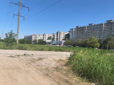 The image depicts a sunny day in a residential area. The foreground is a dirt road with some dry grass and a patch of green vegetation. In the midground, a line of tall, green reeds separates the road from a row of multi-story apartment buildings. These buildings are characteristic of Soviet-era architecture, with a uniform appearance and balconies on each floor. A power line pylon with thick cables stretches across the left side of the frame, silhouetted against a clear blue sky. Beyond the apartment buildings, more trees and some smaller structures are visible. Cars are parked along the street in front of the buildings.
The scene appears to be set in a Russian city, possibly during the daytime due to the bright sunlight and clear sky, suggesting good weather. The overall impression is one of a typical urban landscape on a warm day. No people are explicitly visible in the image. There is some text visible on a building facade in the midground, appearing to be part of a store's signage, but it is too small and blurry to read. The location context provided, Верхнетемерницкий, Russia, aligns with the architectural style of the buildings.