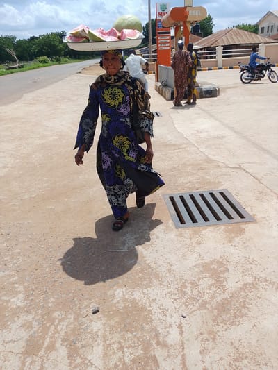 A woman in traditional attire is walking along a paved area, carrying a large tray of sliced watermelon and a whole melon on her head. She is wearing a long, dark blue dress with a colorful, swirling pattern of yellow, purple, and white. Her head is covered with a matching scarf. She is walking from left to right in the frame, with her shadow cast on the ground beside her.

In the background, there is a gas station with an orange and white sign that reads "SIA GLOBAL" and "DIESEL". A few other people are visible at the gas station, including two men in traditional clothing talking near the pumps and a man riding a motorcycle. The scene appears to be outdoors during the daytime, with a bright sky and some clouds. The ground is a mixture of concrete and dirt. The overall setting suggests a rural or semi-urban area in Nigeria.