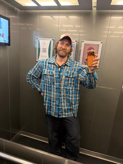 A man is taking a selfie in what appears to be an elevator. He is wearing a blue and yellow plaid shirt, dark pants, and a baseball cap. He is holding an orange phone in his right hand. Behind him are posters and a monitor. The elevator's interior is made of gray panels. The image was taken in Repino, Russia.