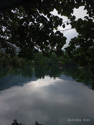A tranquil lake scene is captured in Verkhnyaya Balkariya, Russia, on July 26, 2026, at 15:53. Lush green trees frame the foreground, with their dark silhouettes creating a dappled effect against the bright sky. The calm surface of the lake reflects the surrounding mountains and the cloudy sky, creating a mirror-like image. In the distance, on the far bank of the lake, several buildings are visible, nestled amongst the dense forestation. The overall impression is one of peaceful natural beauty, undisturbed by human activity.