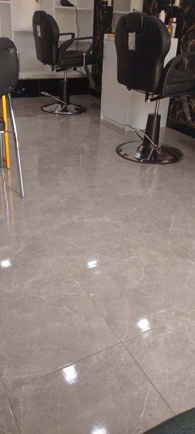The image displays the interior of an unoccupied commercial establishment, identified as being in Matathani ward, Kenya, likely a barbershop or salon. The perspective is low, looking across a clean, polished floor composed of light gray, rectangular tiles with white veining patterns; multiple bright reflections from overhead lighting are visible on the tiled surface. In the midground, two black, cushioned salon-style chairs with chrome hydraulic bases and footrests are present, positioned facing the background. Both chairs are unoccupied. A partial view of a third chair, featuring silver legs and a yellow frame element, is visible in the extreme foreground left. The background reveals white shelving units, a dark, possibly black, wall section with a white marble-like pattern, and a white counter surface holding an assortment of bottles and containers. A black hairdryer is visible in the upper left background, suspended near a shelf. No individuals or active interactions are depicted within the scene.