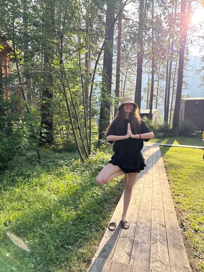In Barangol, Russia, a woman stands in a tree pose on a wooden walkway amidst a forest. She wears a black t-shirt, black shorts, a beige hat, and brown sandals. Her hands are pressed together in a prayer position at her chest. The sun filters through the trees, casting shadows on the ground. To the left of the walkway is lush green grass and trees, while to the right are more trees and some small wooden structures. The background suggests a natural, possibly rural or resort setting.