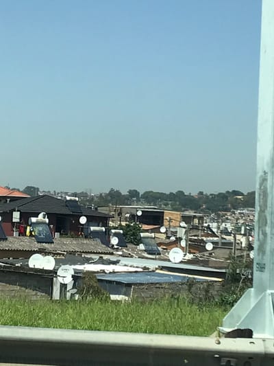 The image depicts a densely populated residential area in Sandton, South Africa, viewed from an elevated perspective under a clear, light blue sky with distant haze. In the immediate foreground, a metal guardrail runs horizontally, partially obscured by green grass and weeds. To the far right, a segment of a light-colored, possibly galvanized steel, utility pole structure is visible.
The midground is filled with numerous single-story residential structures, packed closely together. Roof materials vary, including dark tiles and corrugated metal sheets. A prominent feature across many rooftops are cylindrical solar water heaters, typically accompanied by dark collector panels. Additionally, numerous small, white satellite dishes, some bearing "DStv" or "OSN" branding, are affixed to the roofs and exterior walls of various dwellings. On one dark-tiled roof in the mid-left, clothes are visible hanging on a line. The background shows an undulating landscape with scattered trees and vegetation. No individuals or active human interactions are discernible within the frame.