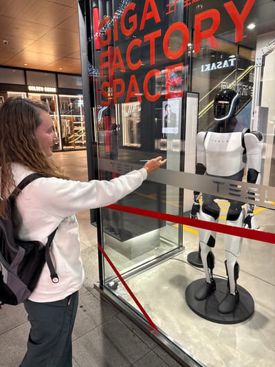 A person with long curly hair, wearing a white sweatshirt and black pants with a black backpack, points their right index finger towards a Tesla Optimus robot displayed in a glass enclosure. The robot is predominantly white with black accents, standing on a circular black base. The display is located in a storefront window with red lettering spelling "GIGA FACTORY SPACE" visible on the glass. The reflection in the window reveals the interior of a clothing store named "GOLDEN GOOSE". The scene is in Jinjiang District, Chengdu, China, indicated by the shop name "TASAKI" written in Chinese characters on the top left corner of the glass enclosure. The ground outside the enclosure is paved with tiles. A red tape stretches across the display.