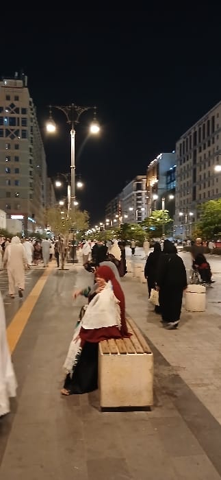The image displays a bustling nighttime scene on a wide, paved pedestrian promenade in Medina, Saudi Arabia. Multi-story modern buildings with numerous illuminated windows flank both sides of the walkway, receding into the distance. Tall, ornate streetlights with multiple glowing globes line the central median, which contains small trees or shrubs.

Numerous individuals populate the scene. In the mid-ground, slightly right of center, a woman is seated on a light-colored stone bench with horizontal wooden slats. She wears a dark dress, a dark red head covering, and a white garment draped over her shoulders. Her bare feet, with sandals, are visible. Most other people are walking, with several men in white thobes and many women in dark abayas and head coverings. A person in a white thobe is partially visible in the left foreground, walking away from the viewer. The ground is composed of dark gray pavers, with a distinct yellow line running along the left edge of the promenade. The overall environment is brightly lit by artificial sources.