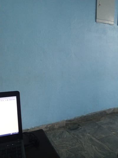 In Bori, Nigeria, a laptop sits on a dark surface in front of a pale blue wall. The laptop screen is illuminated, displaying what appears to be a web browser with a white page. To the upper right of the wall, a light-colored electrical panel is mounted. The floor below the wall is tiled with a dark, marbled pattern. The scene is illuminated by ambient light, creating a slightly dim atmosphere.
