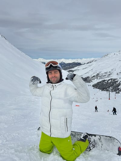 A male individual is depicted kneeling on a snowboard in a snow-covered mountain environment in Les Allues, France. The subject, facing the camera, wears a black helmet with orange-tinted goggles positioned on top, a white quilted puffer jacket with a black central zipper, neon green ski pants, and grey-white gloves. He is flexing both arms at the biceps, forming fists, and smiling. The snowboard is partially visible beneath him. The background features extensive snow-covered mountain terrain. To the left, a steep, smooth snowfield ascends. To the right, a ski slope with several skiers and snowboarders, appearing as dark figures, and multiple red ski lift poles are visible. Distant snow-capped mountain ranges stretch across the horizon under an overcast sky.