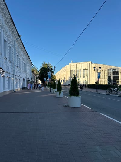The image depicts a street scene in Yazvitsevo, Russia. On the left, a white building with ornate architectural details lines the sidewalk. Several people are walking on the sidewalk in front of the building. Across the street, a large, light-colored building with a classical facade stands under a clear blue sky. Planters with conical evergreen trees are placed along the sidewalk in front of this building. A road runs alongside the sidewalk, and there are bicycles parked near a modern glass-enclosed structure. Power lines are visible stretching across the sky. The overall environment suggests a daytime scene in a city.