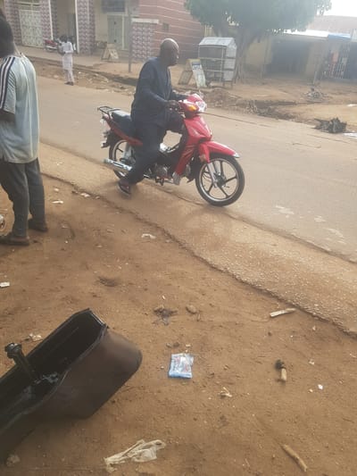 The image captures an outdoor street scene in Tukur Tukur, Nigeria, under bright daylight. The main subjects are three men, a red motorcycle, and a large black pan.
In the midground, a bald man wearing a dark blue long-sleeved shirt and trousers is seated on a red Honda motorcycle, which is stationary on a paved road. His feet are on the ground, suggesting he is either paused or about to move. The motorcycle faces towards the left side of the frame (if upright).
In the foreground, partially visible on the bottom right (if upright), is another man wearing a light-colored shirt with striped sleeves and dark trousers, standing on the dirt shoulder. Further in the background, a third man in white attire is seen standing near the road.
The immediate foreground, to the right of the paved road (if upright), consists of reddish-brown dirt scattered with small stones, pieces of wood, and various items of litter, including plastic bags. A prominent object in this area is a large, black, rectangular pan, possibly a vehicle oil pan, lying on the dirt.
The background features several modest buildings with corrugated iron roofs, indicative of local architecture. A small, simple kiosk or stall is visible near the buildings, with faint text that appears to read "POST" on a blue sign. Trees and sparse vegetation are also visible further back, beyond the buildings. The overall environment suggests a casual, perhaps busy, street in a residential or commercial area.