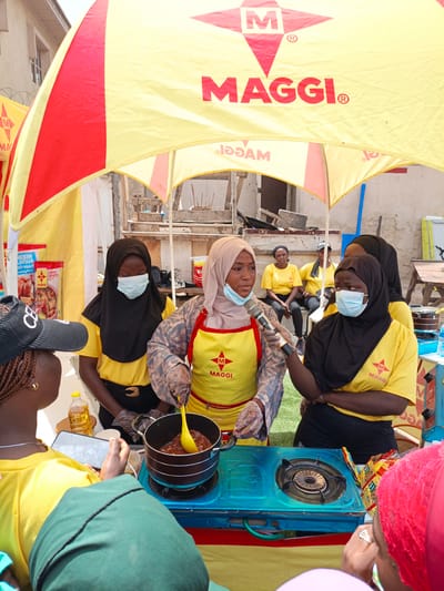A medium shot captures an outdoor cooking demonstration event, likely in Tarauni, Nigeria, under a red and yellow branded canopy displaying "MAGGI." In the foreground, three young women are the primary subjects. On the left, a woman wearing a yellow shirt and a black cap with braided hair and gold earrings is partially visible, holding a smartphone. In the center, a woman in a black hijab and a yellow shirt, wearing a light blue surgical mask, is looking down at a pot on a blue gas stove. She is stirring a reddish sauce with a yellow spoon, wearing clear plastic gloves. To her right, a woman in a light brown hijab and a yellow apron over a patterned long-sleeved shirt, also wearing a blue surgical mask, is speaking into a microphone held by the central figure. In the background, other individuals, some wearing yellow shirts with the Maggi logo, are seated or standing. The environment appears to be an urban setting with buildings visible behind the canopy and some construction materials in the background. Several Maggi product packages are displayed on shelves behind the cooking area. The overall scene depicts a promotional event or a cooking class focused on Maggi products.
