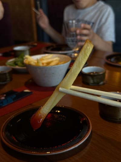A close-up shot shows a single French fry being dipped into a dark liquid in a small, shallow bowl. The French fry is golden brown and has a dollop of red sauce on its tip. Chopsticks are holding the fry. The bowl is dark and circular with a light-colored rim. In the background, out of focus, are other bowls of food, drinks, and people. The scene appears to be in a restaurant or dining setting. The location is Piņķi, Latvia.