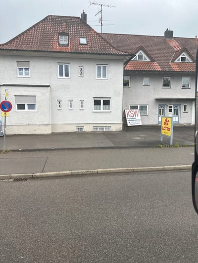 The image shows a street view in Memmingen, Germany, featuring a multi-story building with a reddish-brown tiled roof. The building has several windows, some with white frames and others with decorative metal grates. A white and red A-frame sign is visible in front of the building, displaying the text "KSW" and information about "Kfz-Prüfstelle Kfz-Sachverständige" along with a phone number and address. To the right of this sign, a yellow vertical sign with a red cloverleaf logo and "LOTTO Bayern" is also present. The street in the foreground appears to be asphalt, with a concrete curb and a drain grate visible. The sky is overcast.