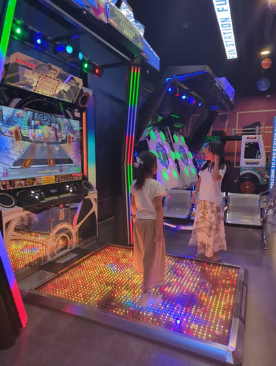 Two young girls are playing a dance arcade game in a dimly lit entertainment venue, likely a game center. The game console features a large screen displaying a rhythm-based game with glowing graphics, and a lighted dance pad on the floor. The dance pad is illuminated with a vibrant array of red, yellow, blue, and purple LEDs. One girl, facing away from the camera, wears a light-colored t-shirt and tan pants, with white sneakers. The other girl, facing the first, is in a white t-shirt and a long, patterned skirt, with white sandals. The arcade machine itself is branded with "KONAMI" and "DANCERUSH STARDOM" logos. In the background, more arcade machines are visible, along with seating and decorative planetary models suspended from the ceiling. A sign in the upper right corner reads "FUN STATION FU". The overall environment suggests a modern arcade designed for interactive gaming experiences.