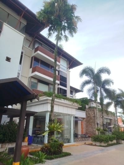 The image captures a contemporary building with a modern architectural style, featuring clean lines, white facades, and extensive use of glass. The building is partially obscured by lush tropical vegetation, including several tall palm trees that reach towards a pale, overcast sky. The foreground shows a paved walkway with landscaping that includes various green plants and a large terracotta pot. On the right side of the image, a stone-clad building with large windows is visible, also surrounded by greenery. Outdoor seating areas with tables and chairs are set up in front of a glass-enclosed space, suggesting a commercial establishment like a cafe or restaurant. The overall impression is one of a serene, well-maintained outdoor area within a developed, likely tropical, urban environment, possibly a resort or a commercial complex in Ko Kaeo, Thailand. There are no people visible in the image.