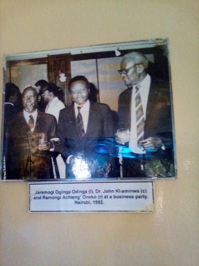 A framed photograph, mounted on a light-colored wall, depicts three adult males at what the accompanying caption identifies as a business party in Nairobi, 1992. The photograph itself shows signs of wear and is covered by a reflective surface, causing a blue glare across the lower half.
From left to right:
1. **Jaramogi Oginga Odinga (l):** A man with dark, short hair, wearing a dark suit, light shirt, and dark tie. He holds a clear glass with a beverage in his right hand and smiles towards the viewer.
2. **Dr. John Khaminwa (c):** Positioned centrally, slightly behind Odinga. He wears a dark suit, light shirt, and a striped tie, smiling towards the viewer. His hair is dark and short.
3. **Ramongi Achieng' Oneko (r):** An older man with gray hair and eyeglasses, wearing a dark suit, white shirt, and a diagonally striped tie. He holds a clear glass with a beverage in his left hand and looks towards the left, displaying a slight smile.
Additional blurred figures are visible in the background, suggesting an indoor social setting. The subjects are formally dressed, consistent with a business event. The white rectangular caption card below the photograph reads: "Jaramogi Oginga Odinga (l), Dr. John Khaminwa (c) and Ramongi Achieng' Oneko (r) at a business party. Nairobi, 1992." The physical location of the image itself is a wall, with no specific address information provided in the image or caption for its current display.