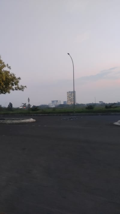 This is a vertical, eye-level, outdoor, daylight photograph taken at Pusakarakyat, Indonesia. The image captures a somewhat hazy sky with a pale grey hue, suggesting it might be early morning or late afternoon. A single, tall, curved lamppost stands prominently in the middle ground, its dark form silhouetted against the sky.

In the background, several modern buildings rise from a landscape of green fields and scattered trees. The most striking structure is a tall, rectangular building with a facade covered in a grid of illuminated windows, giving it a distinctive, patterned appearance. Other buildings, appearing shorter and less distinct, are also visible. The overall impression is that of an urban or semi-urban environment, possibly a developing area or a business district.

In the foreground, a dark asphalt surface, likely a road or parking lot, dominates the lower half of the frame. A circular concrete planter with white markings is visible on the left side of the asphalt. A tree with green foliage is present in the upper left corner, its branches reaching into the frame. The scene appears quiet and still, with no people or vehicles immediately visible, contributing to a sense of calm or even slight desolation. The atmosphere is subdued due to the hazy sky. There is no visible text in the image.