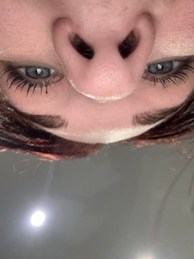 This media file is a close-up, rotated photograph of a person's face, focusing on their nose and eyes. The subjects are a person with fair skin, dark hair, and light-colored eyes. The activity appears to be a casual portrait, as the person is looking towards the camera, but with a slight upward tilt of their head.

Notable details include the person's eyelashes, which are dark and appear to be coated with mascara. The skin texture is visible, showing pores and a slight sheen. The lighting in the image is somewhat diffused, with a bright, circular light source visible in the background, suggesting an indoor setting with artificial lighting, possibly a room with a lamp or overhead light. The background is a plain, gray, slightly textured surface, hinting at a wall or ceiling. There is no visible text in the image. The overall mood of the image is neutral, with no strong emotional cues conveyed. The location context provided is Pervouralsk, Russia, however, there are no visual cues in the image to confirm or suggest this specific location.