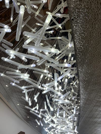 The image shows a close-up view of a modern chandelier made of numerous clear glass tubes containing LED lights. These tubes are suspended by thin wires, creating a complex and layered effect. The lights are illuminated, casting a bright, cool glow. The background is a mix of reflective surfaces, including what appears to be a metallic wall or ceiling with a textured pattern. Some darker areas suggest reflections of the environment above and around the chandelier, possibly including hints of people or other architectural elements. The overall impression is one of contemporary, artistic interior design. The image was captured in Lin'an District, China, though no specific location within the district is identifiable from the visual information provided.